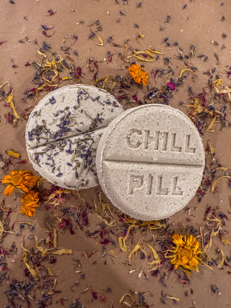 Two bath bombs labeled 'CHILL PILL' on a textured surface with dried flowers.