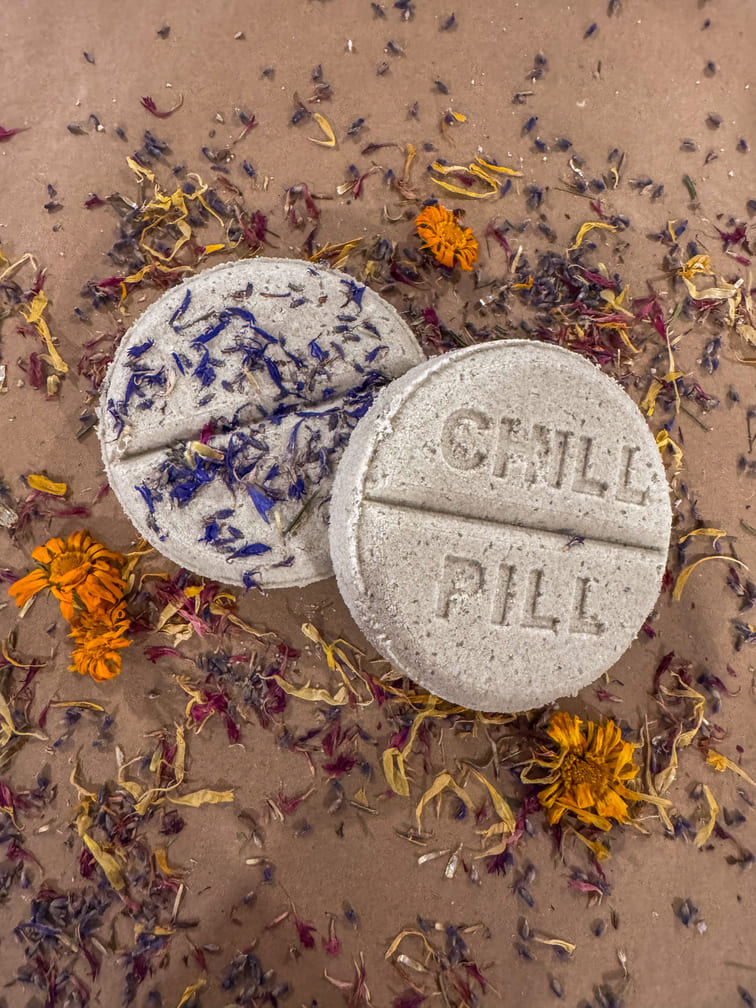 Two bath bombs labeled 'CHILL PILL' surrounded by dried flowers on a brown background
