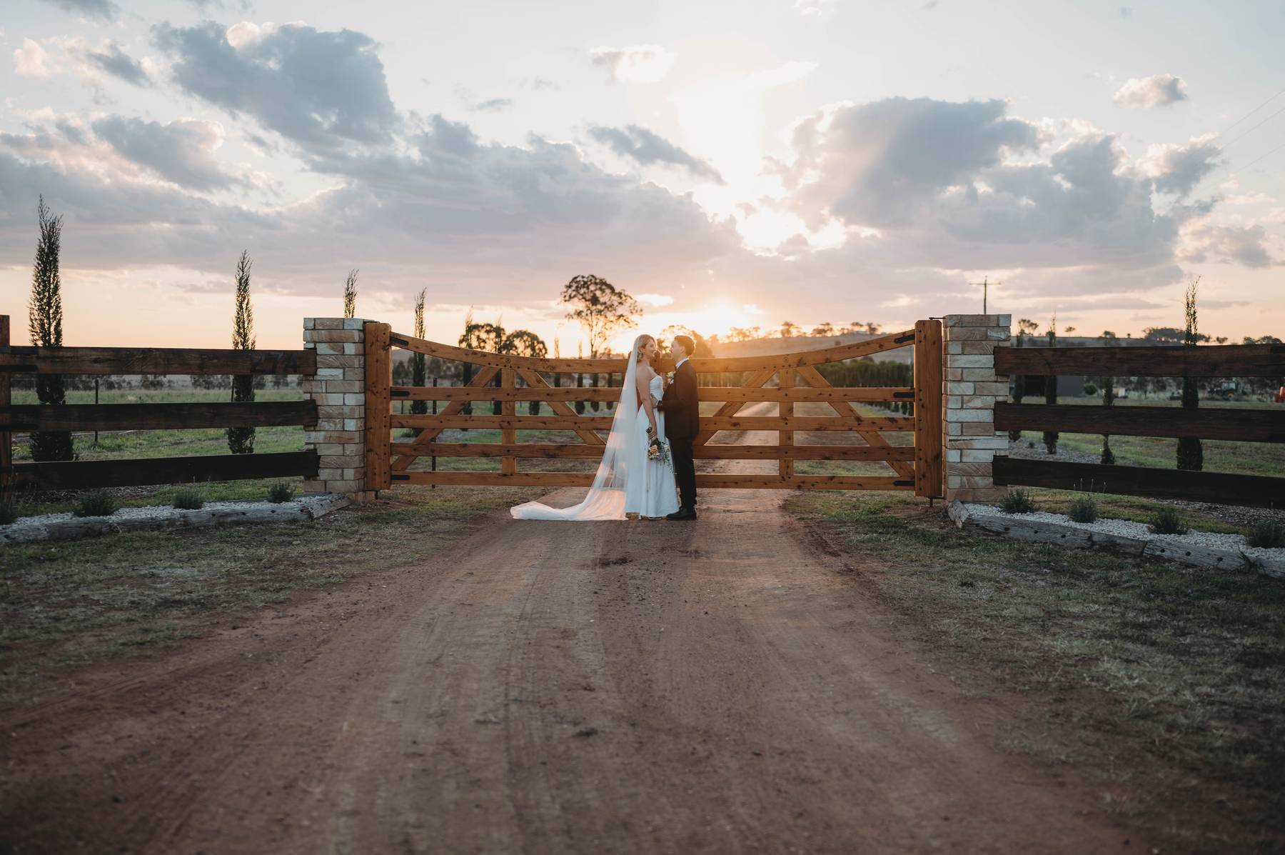 Wedding couple standing in front of a wooden gate with a sunset in the background