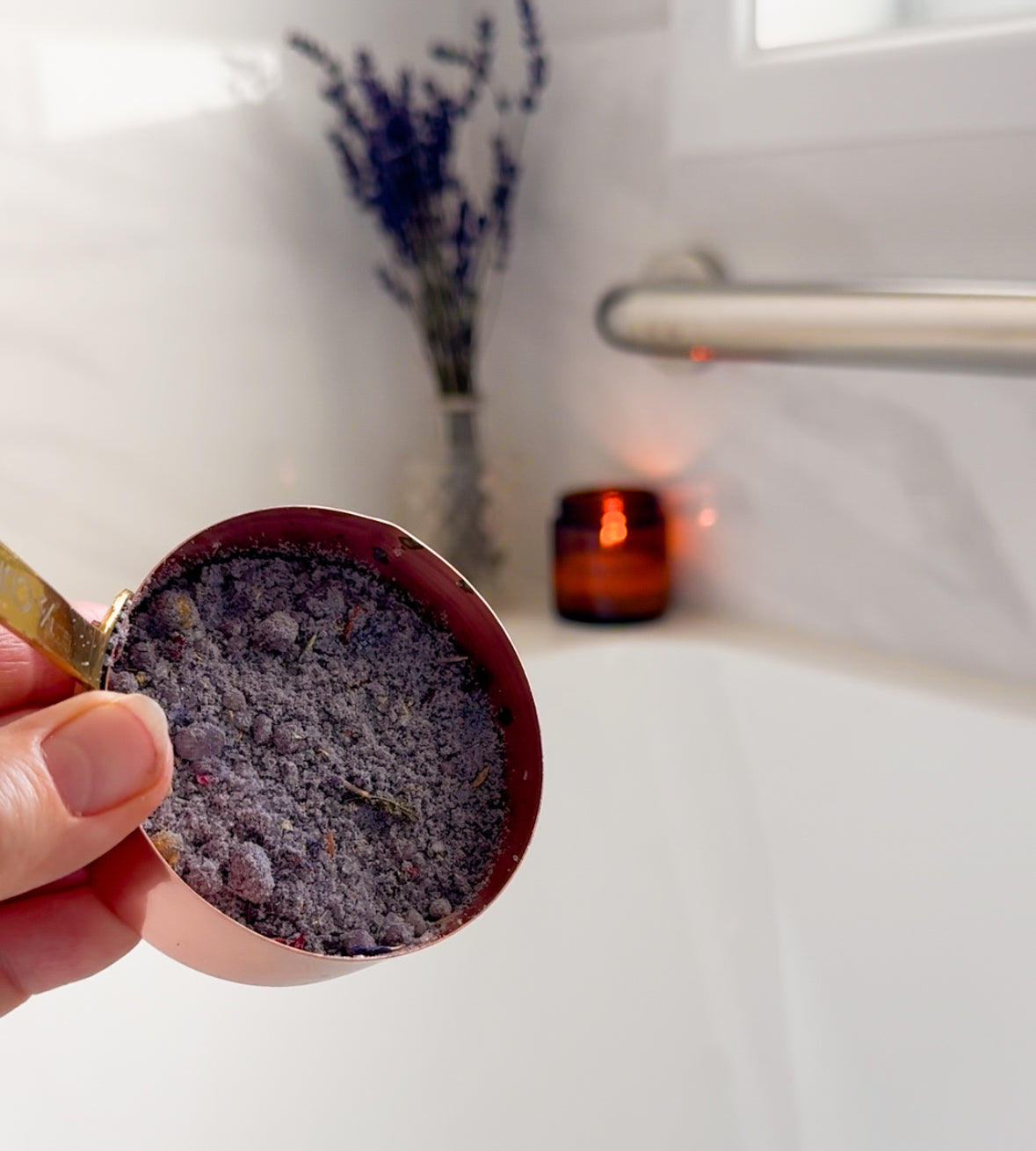 Hand holding a small container of purple bath milk with a bathtub and candle in the background.