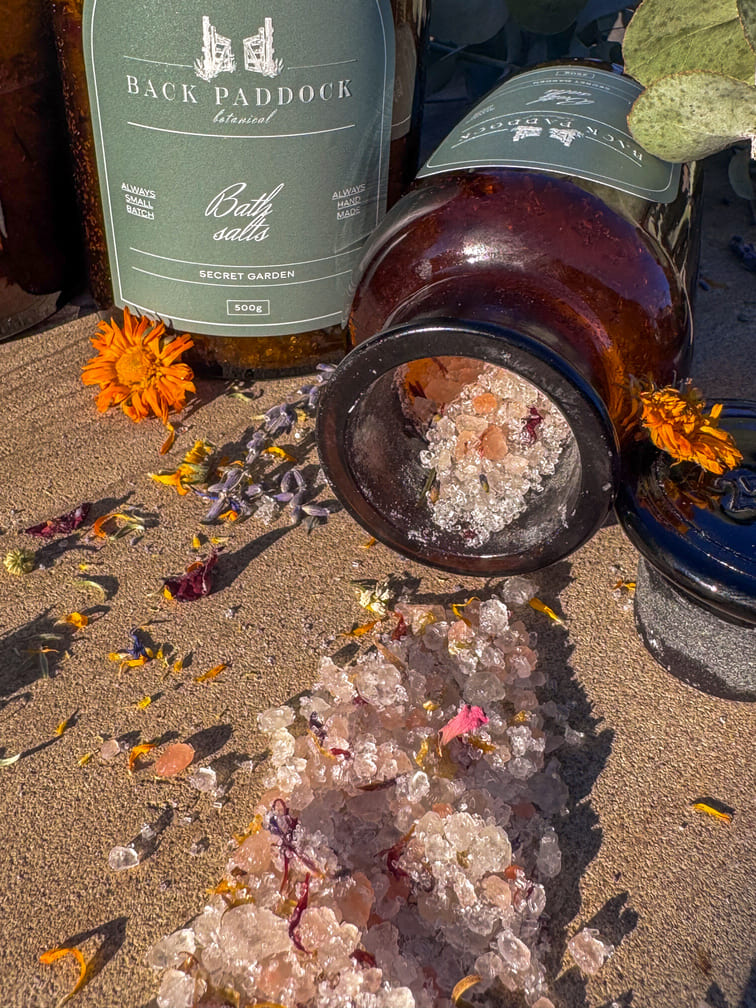 Bottle of Back Paddock Bath salts with a small container of salt on a sandy surface with flowers.