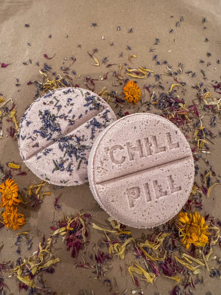 Two bath bombs labeled 'Chill Pill' on a sandy surface with dried flowers.
