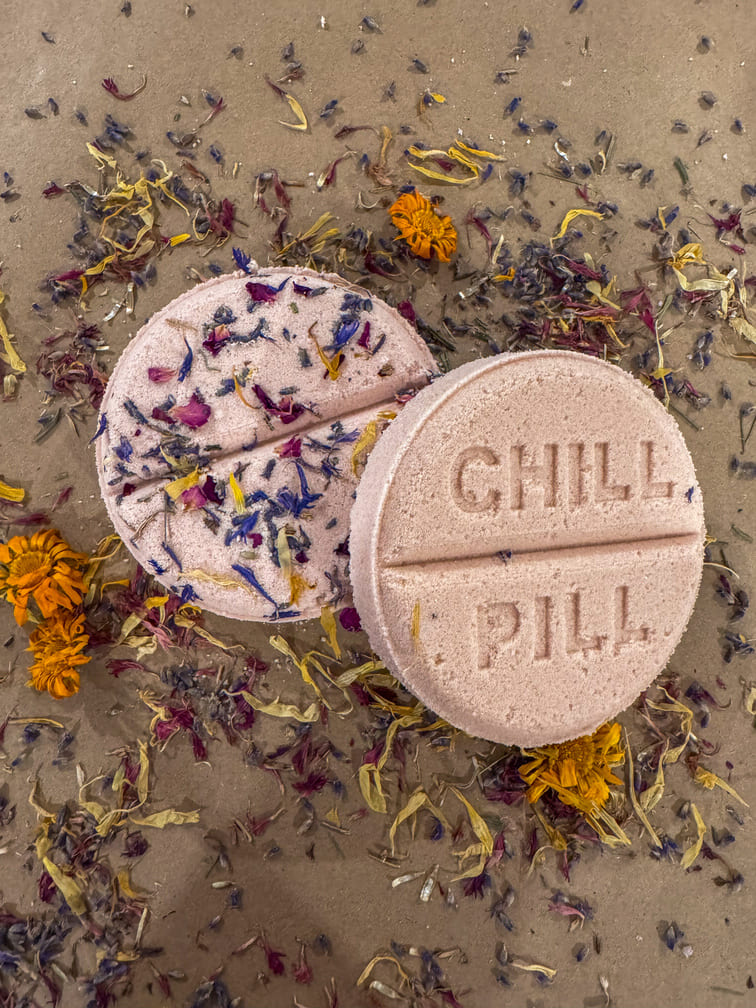 Two round pink bath bombs with 'CHILL PIL' text on a sandy surface with dried flowers.