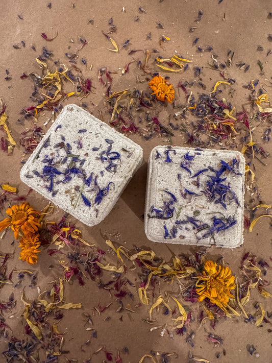 Two cube bath bombs topped with dried herbs on a sandy dried flower covered surface.