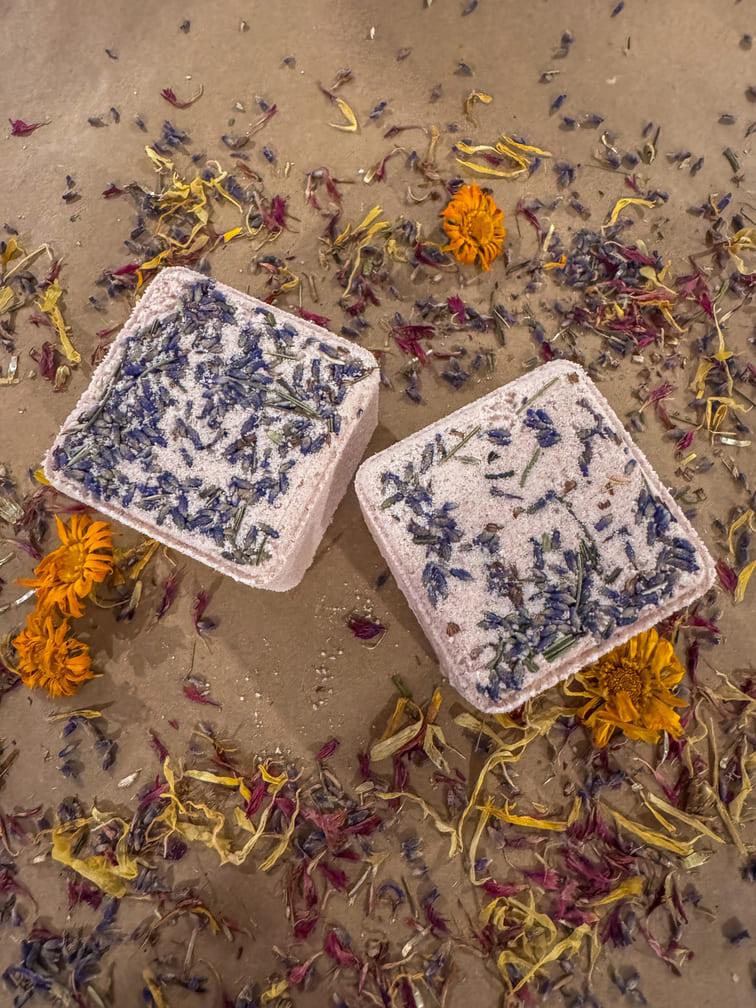 Two cube bath bombs with floral embedded ingredients on a sandy background with scattered flowers.
