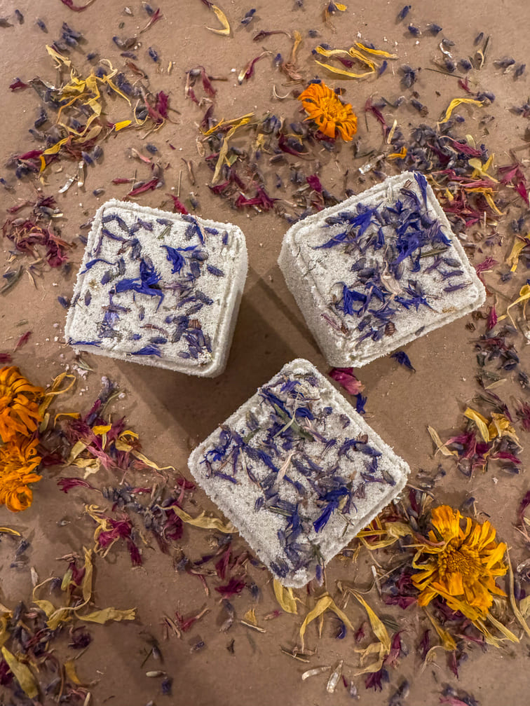 Three cube bath bombs with floral petals and flowers on a sandy background