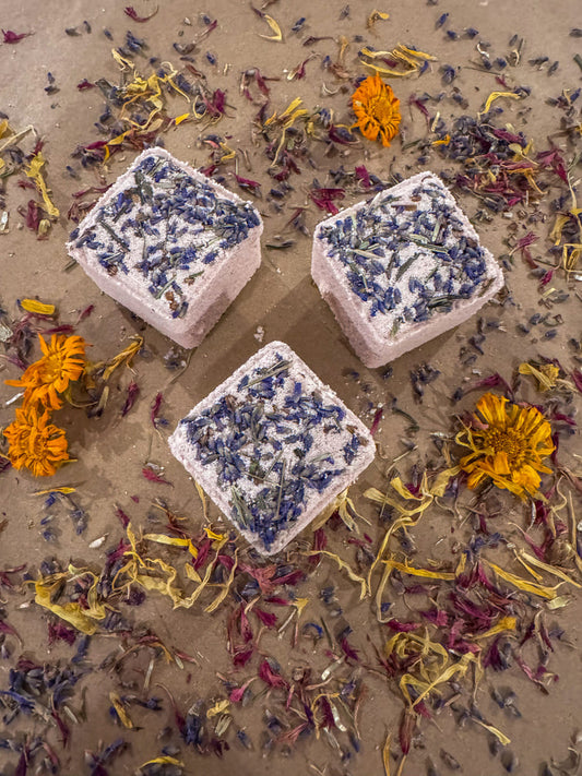 Three cube bath bombs with dried flowers and herbs on a sandy background