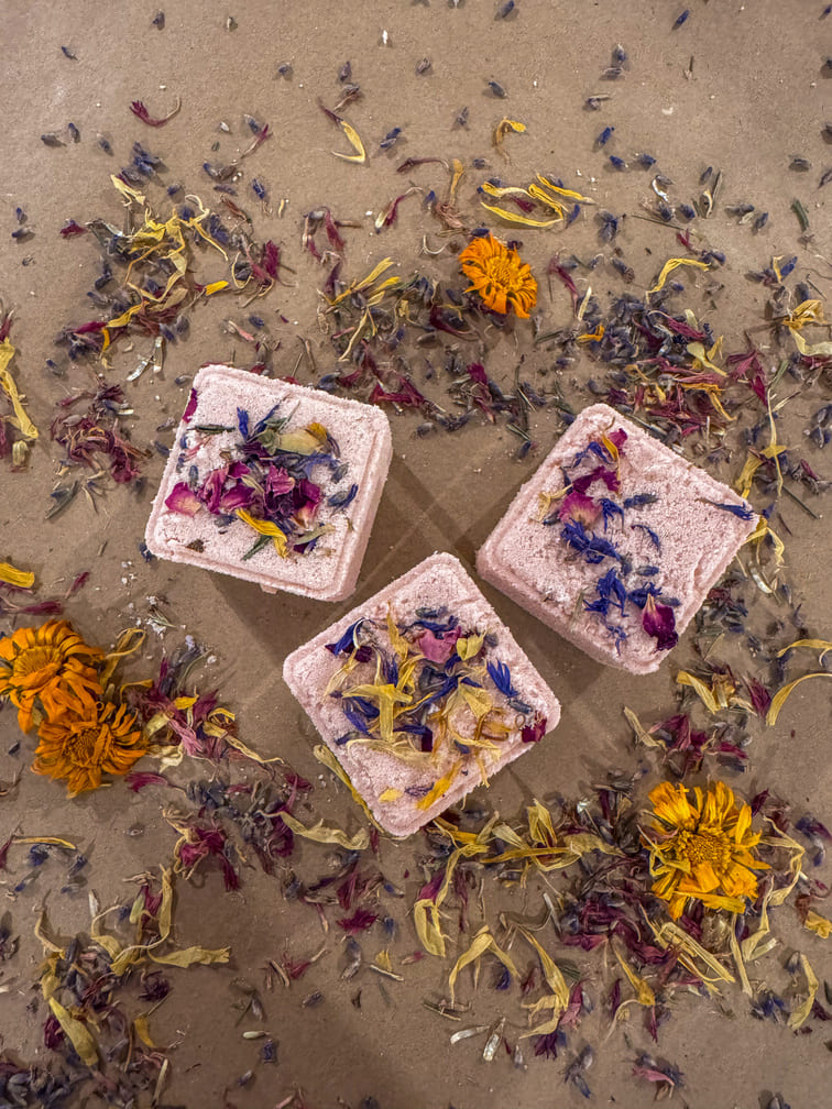 Three square bath bombs with floral petals on a sandy surface with scattered flowers.
