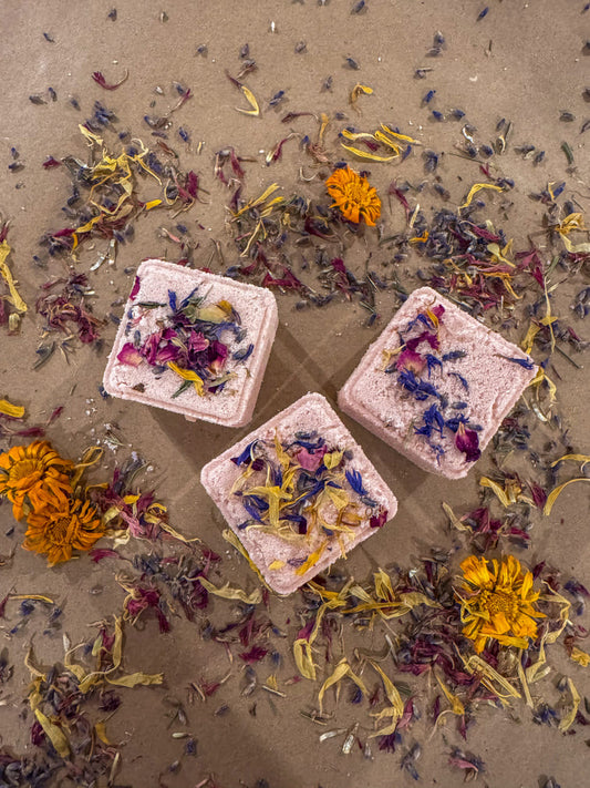 Three square bath bombs with floral petals on a sandy surface with scattered flowers.