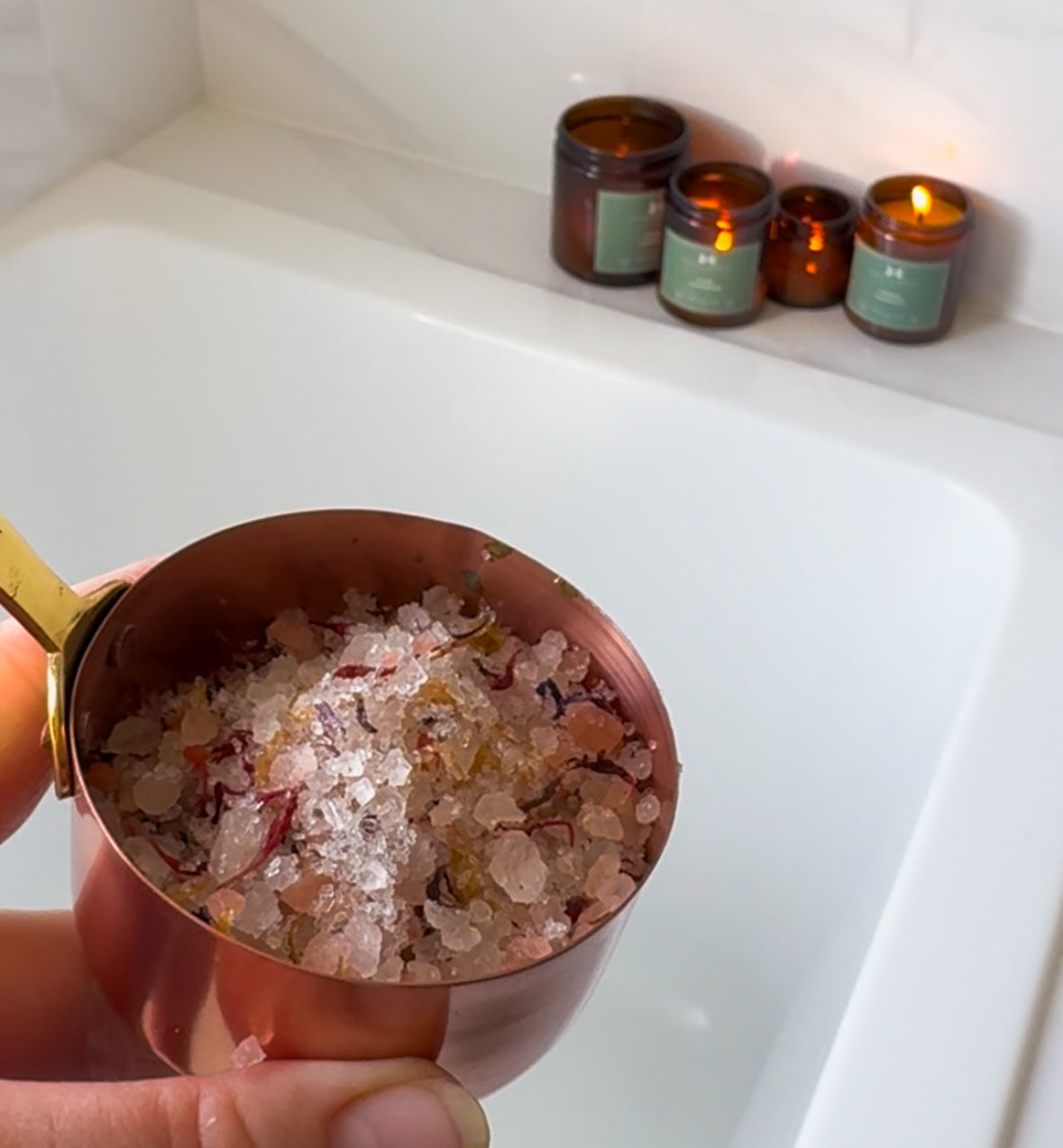 Copper measuring cup filled with bath salts held over a bathtub with lit candles in the background.