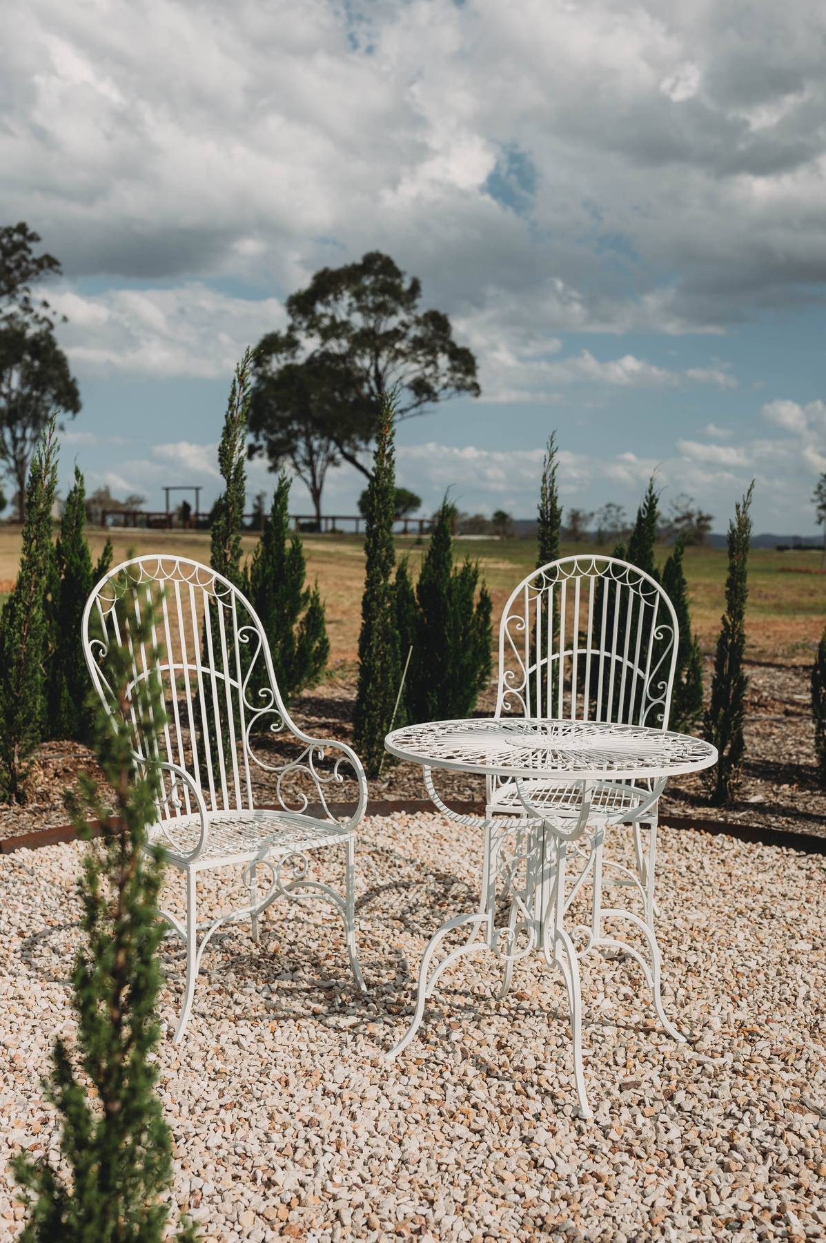 White metal chairs and table set on a gravel area with trees and cloudy sky in the background