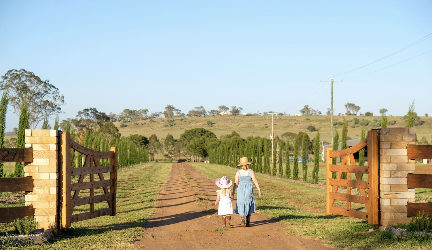 Two children walking along a dirt path with wooden gates on either side, set against a scenic landscape.