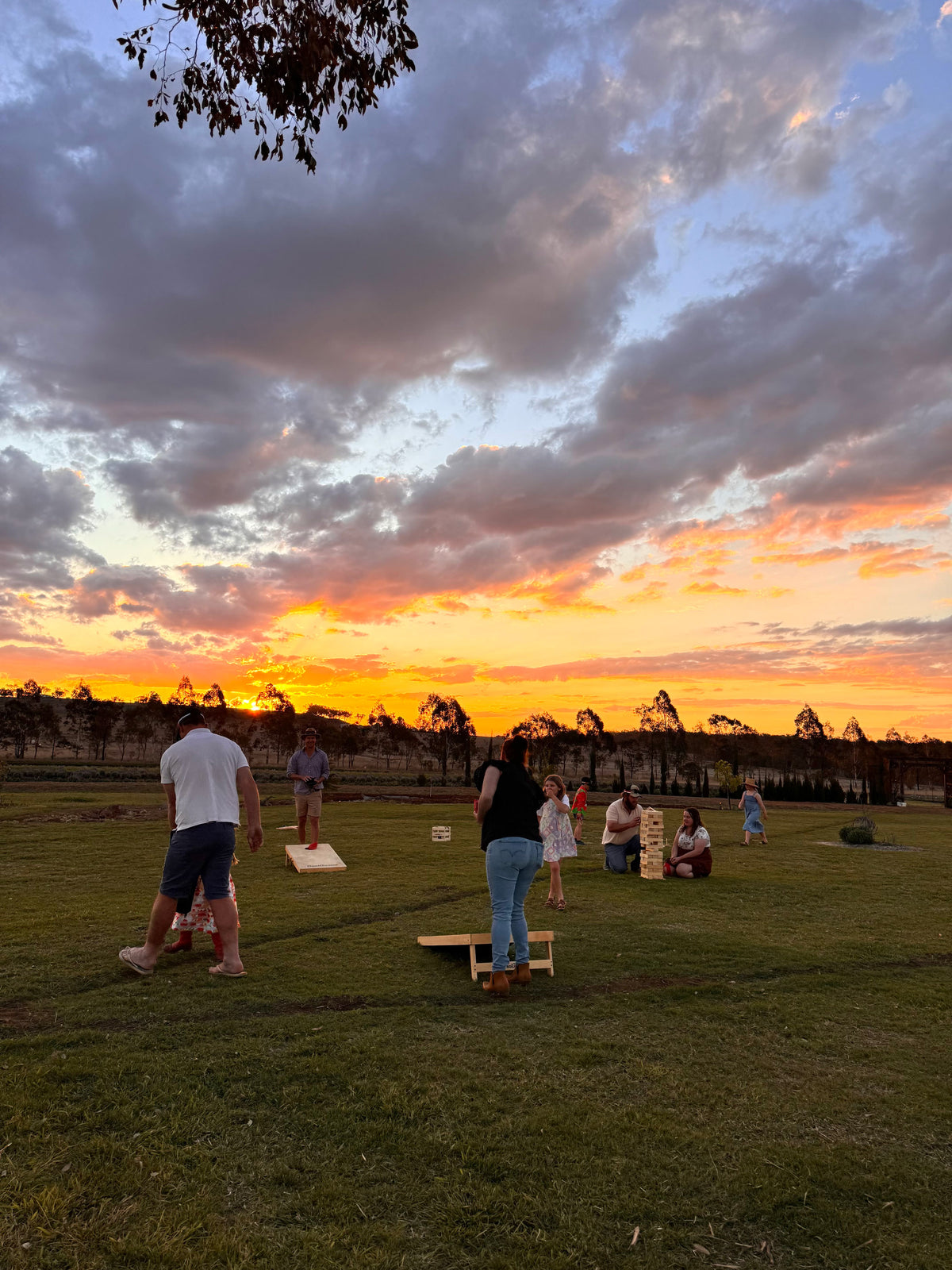A group of people playing cornhole on a grassy lawn at sunset, with a dramatic orange and blue sky filled with clouds behind them.