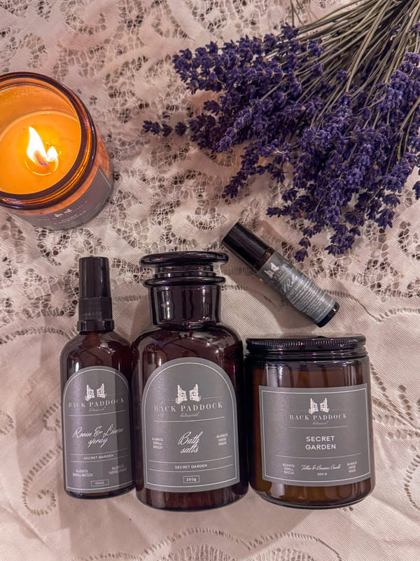 Three dark glass bottles of Back Paddock Botanical Products with labels, a lit candle, and lavender flowers on a lace surface.