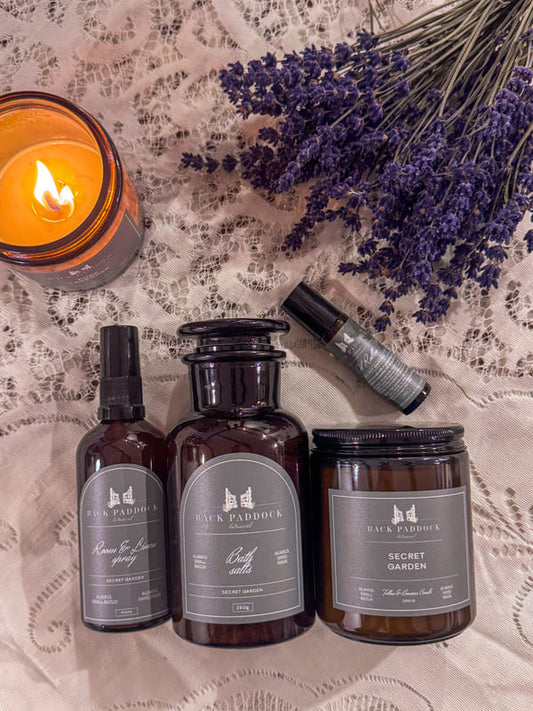 Three dark glass bottles of Back Paddock Botanical Products with labels, a lit candle, and lavender flowers on a lace surface.