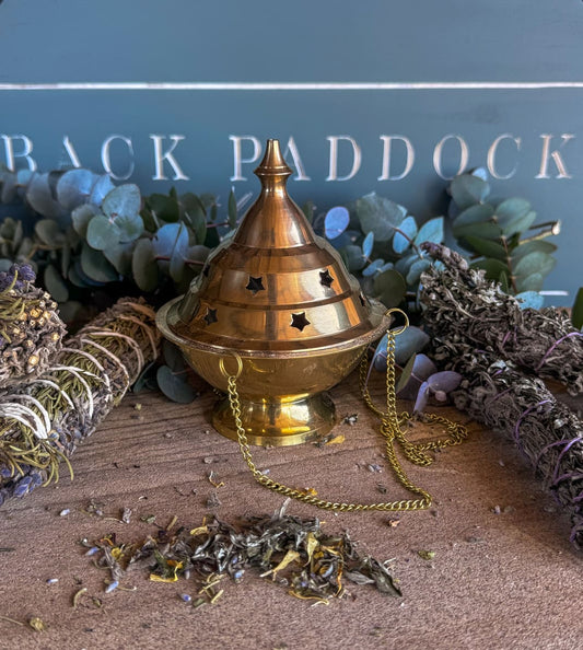 Incense burner with chain on a wooden surface with herbs and eucalyptus leaves.