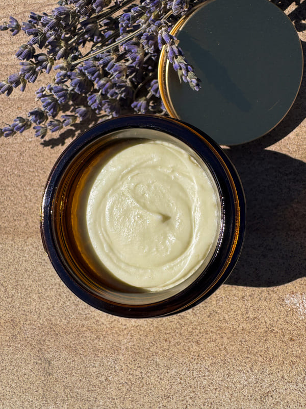 Jar of lemon myrtle tallow cream with a gold lid on a textured surface with lavender flowers.