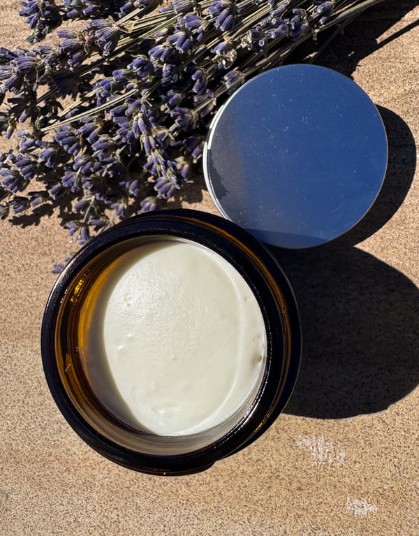 Open jar of tallow cream with a silver lid on a sandy surface with lavender flowers.