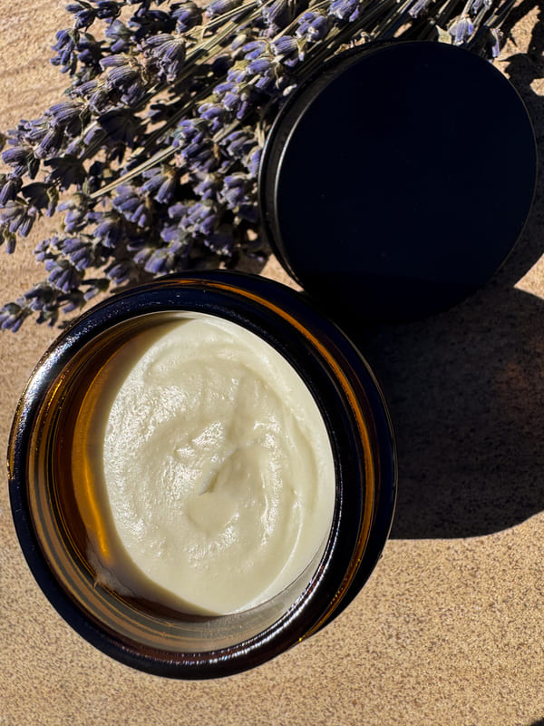 Open jar of tallow cream with a black lid on a sandy surface, surrounded by lavender flowers.